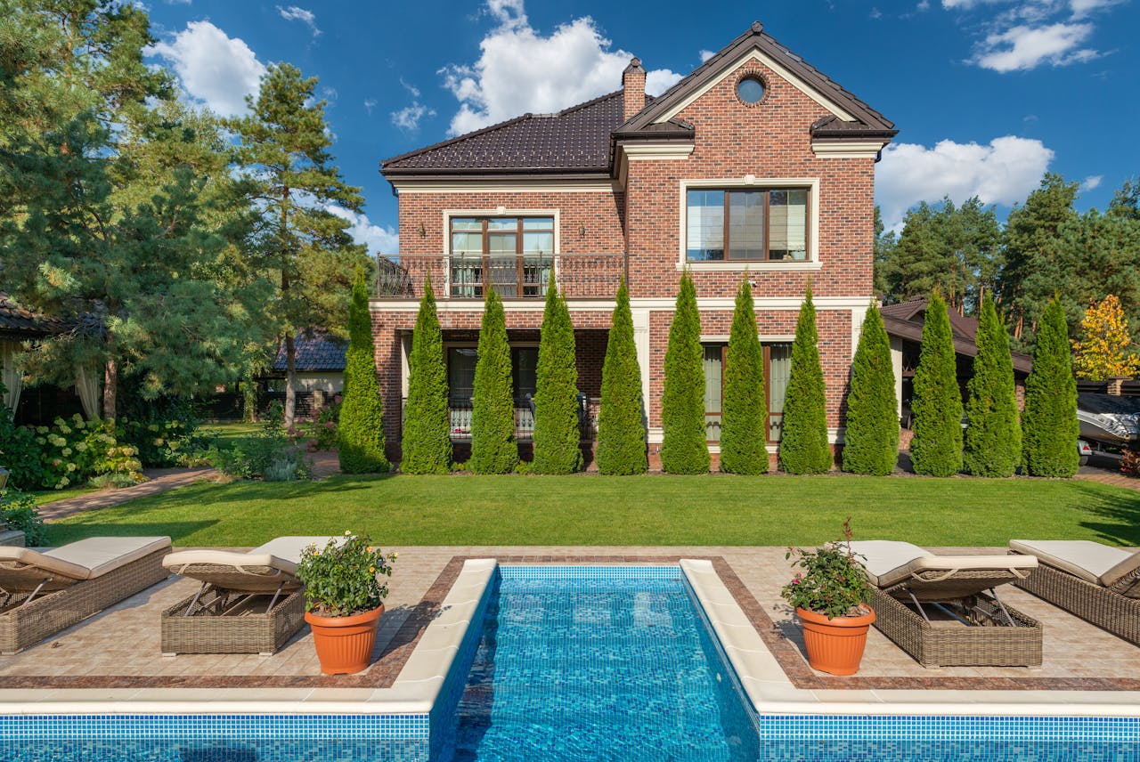 Facade of brick dwell villa with green bushes on lawn and sunbeds near swimming pool